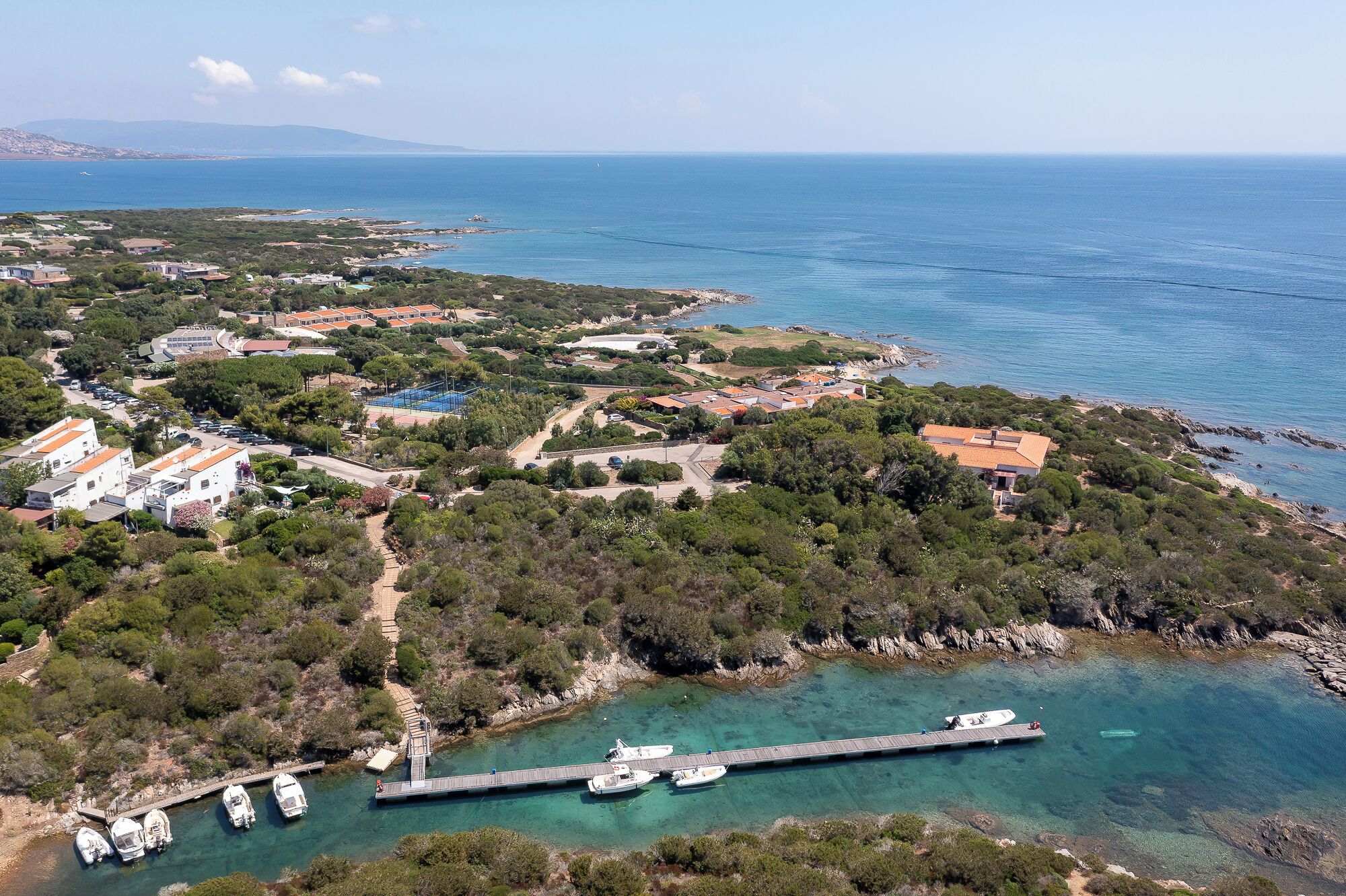 Vista aérea de una zona costera con edificios residenciales, un muelle con barcos y aguas azules cristalinas.
