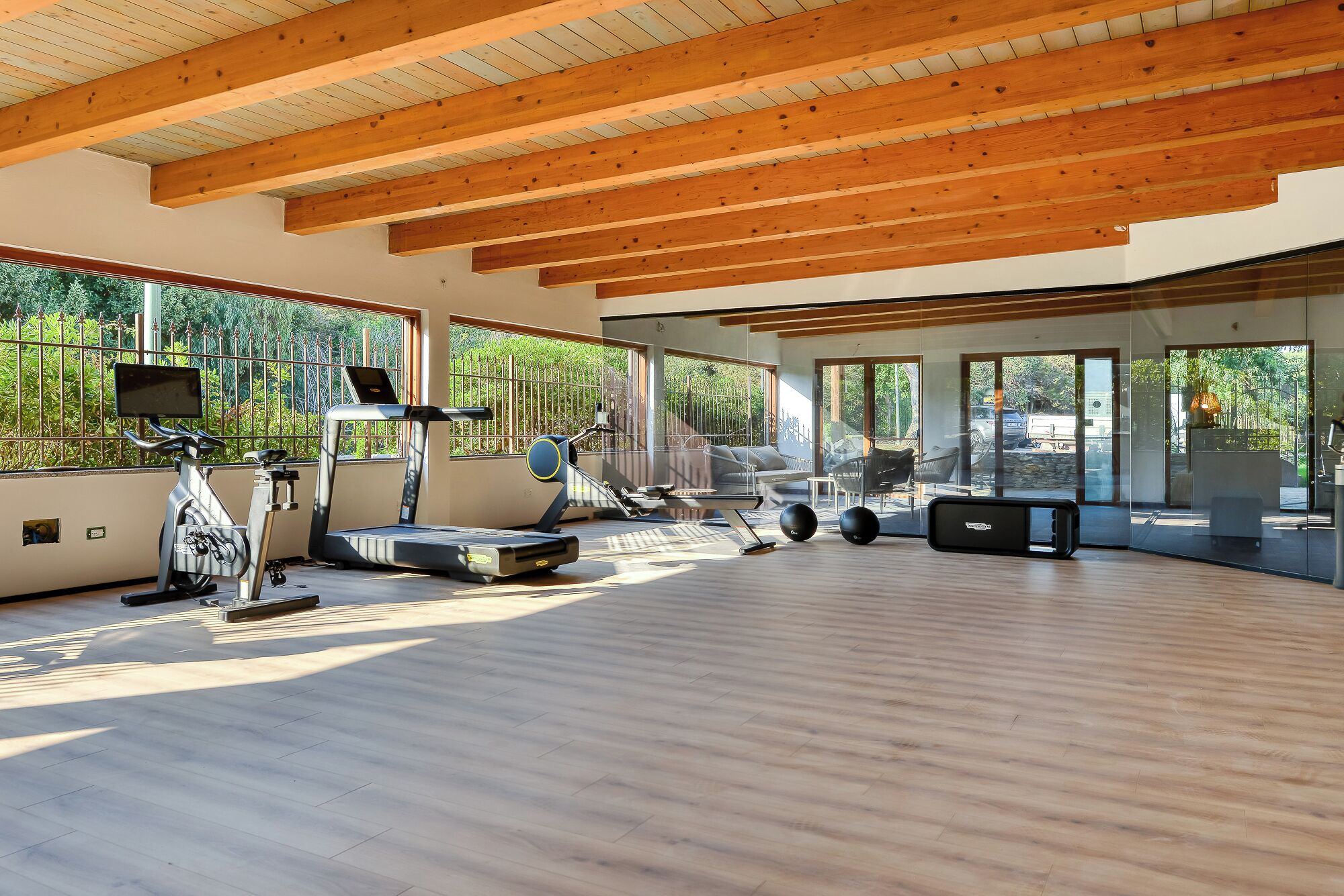 Ein geräumiger Fitnessraum mit Holzbalken, Trainingsgeräten und großen Fenstern, die einen Blick ins Grün ermöglichen.