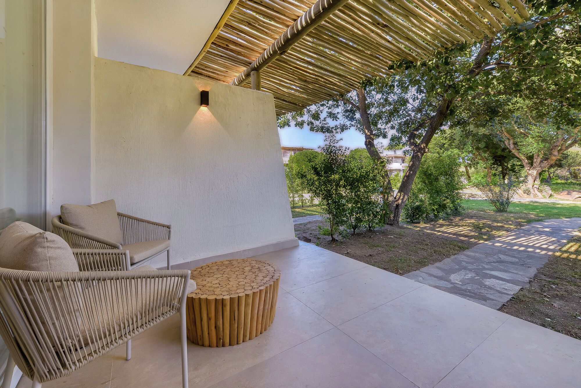 A covered outdoor seating area with wicker furniture and a round wooden table, overlooking a landscaped garden with trees and a pathway.