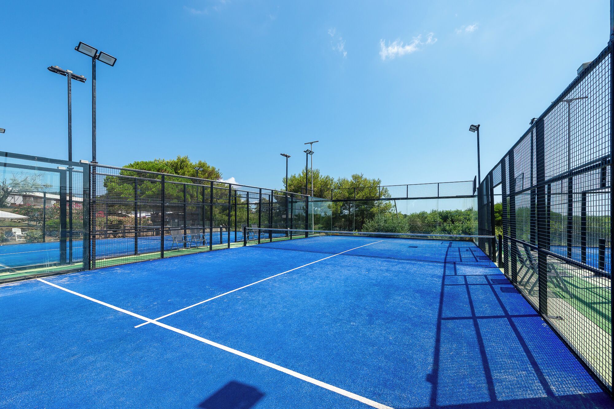 An outdoor tennis court with a blue surface, surrounded by a black fence, and equipped with overhead lighting.