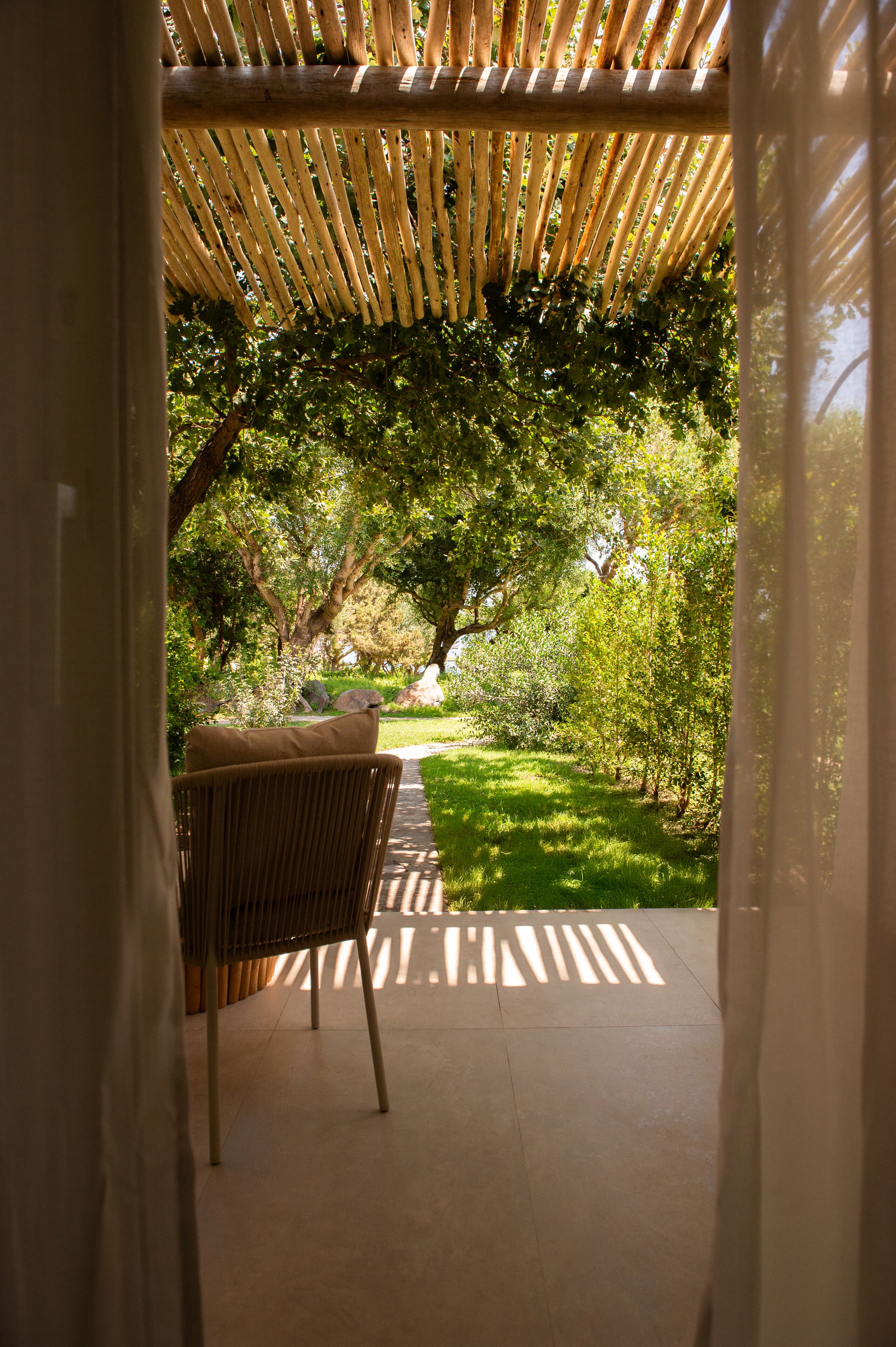 A covered outdoor seating area with a wicker chair and a cushion, surrounded by lush greenery and trees.