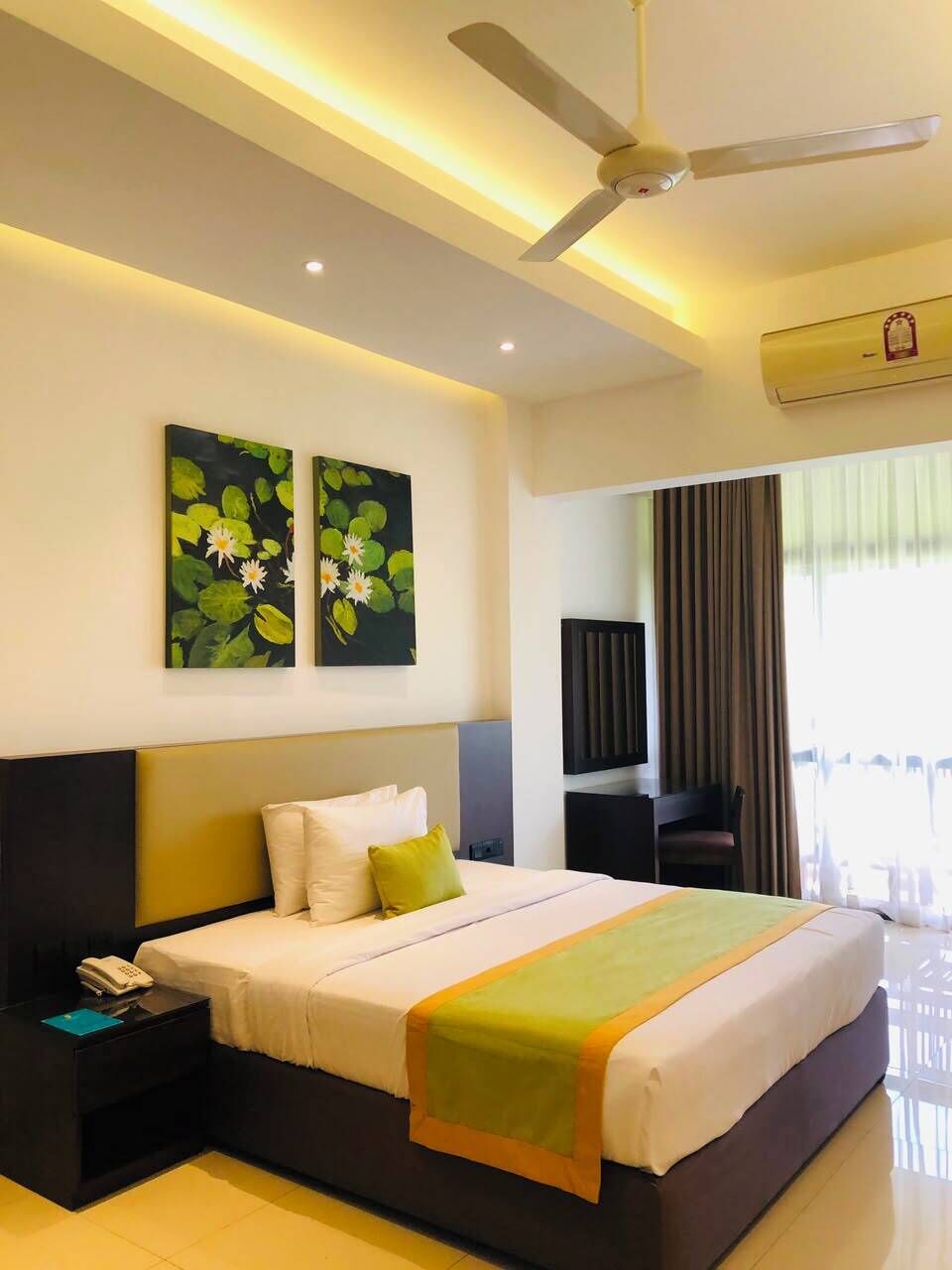 Une chambre d’hôtel comprenant un lit, une table de chevet, un ventilateur de plafond, des œuvres d’art et une fenêtre avec des rideaux.