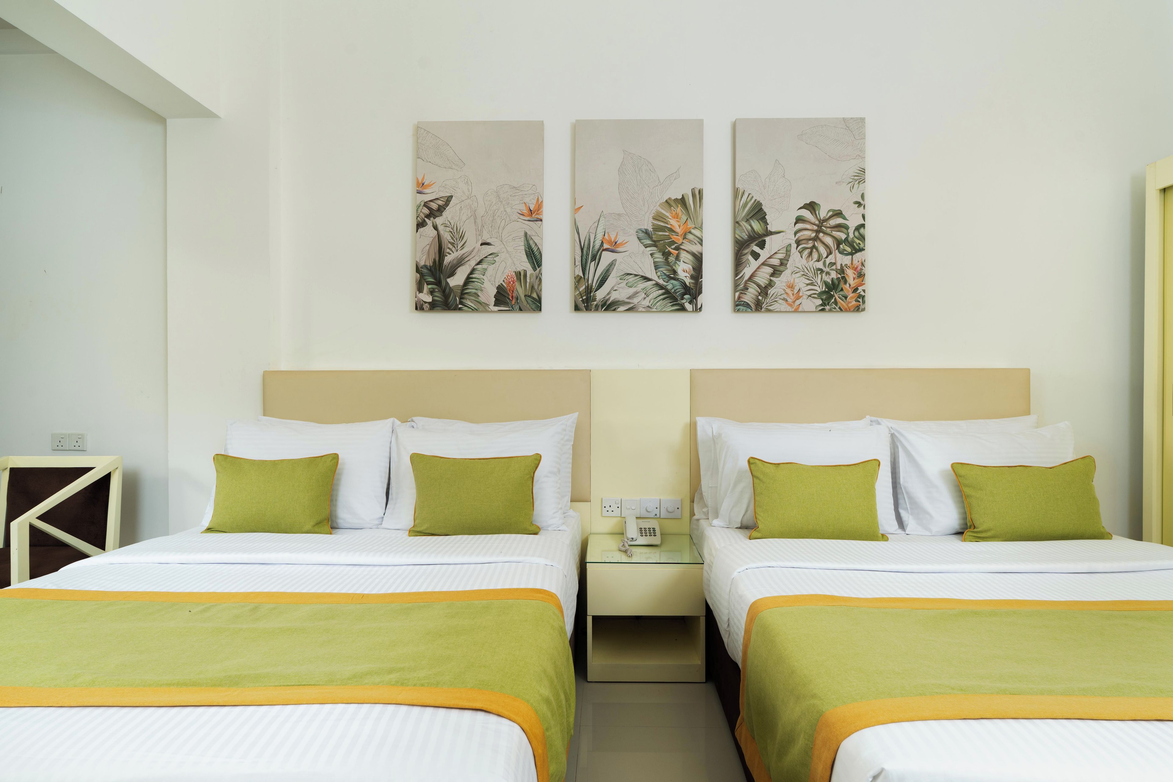A hotel room with two beds, each with green pillows and a green throw blanket. There are two nightstands with a phone and a lamp. The walls have three botanical artwork pieces.