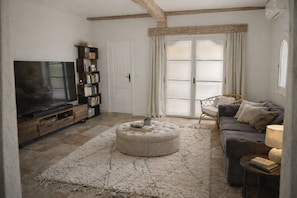 A living room with a television, bookshelf, ottoman, sofa, and a large window with curtains.