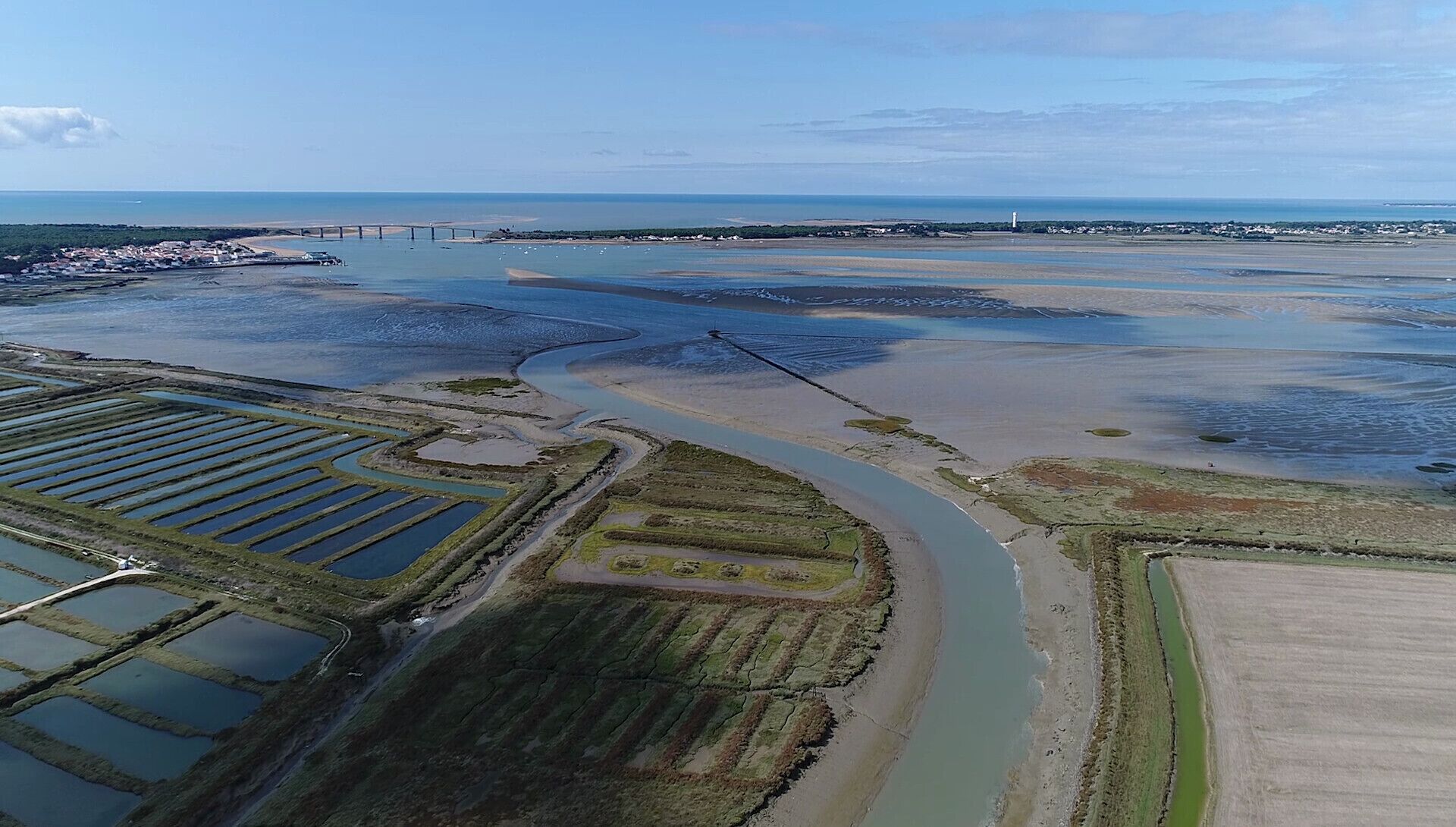 An aerial view of a coastal area with a river, a bridge, and a lighthouse in the distance.