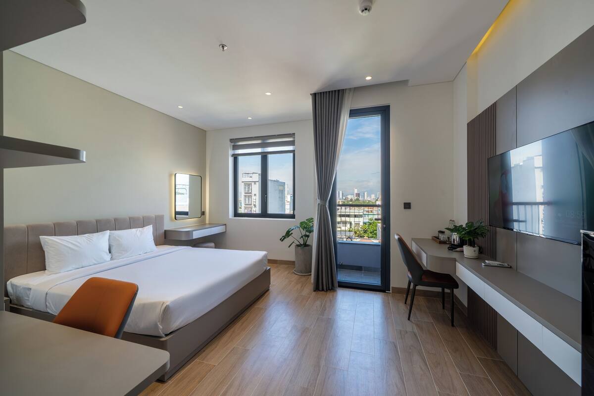Habitación de hotel moderna con una cama grande, un escritorio, una silla, un televisor y un balcón con vistas a la ciudad.