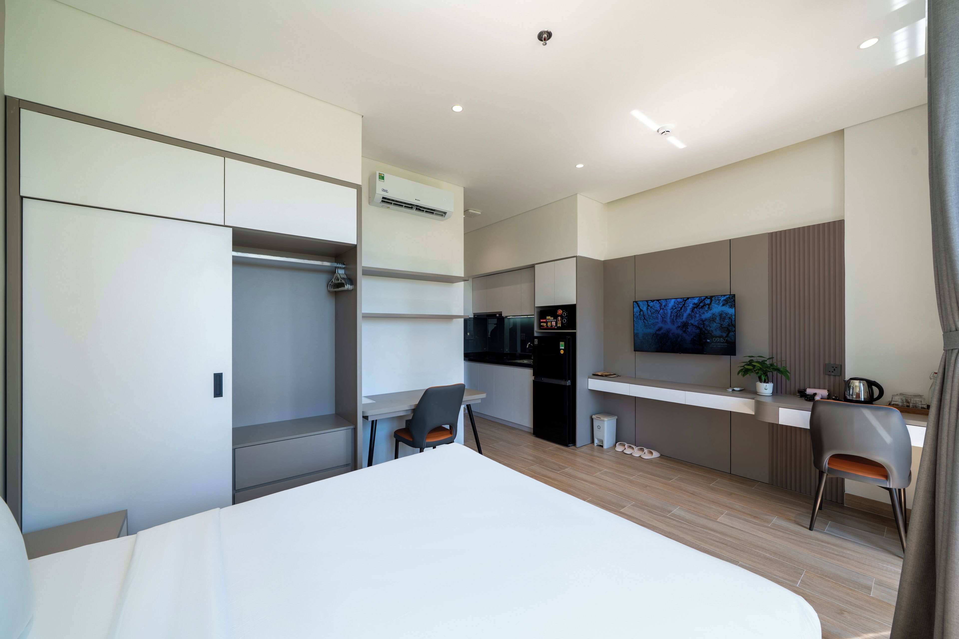 A modern hotel room with a bed, a desk, a chair, a TV, and a kitchenette.