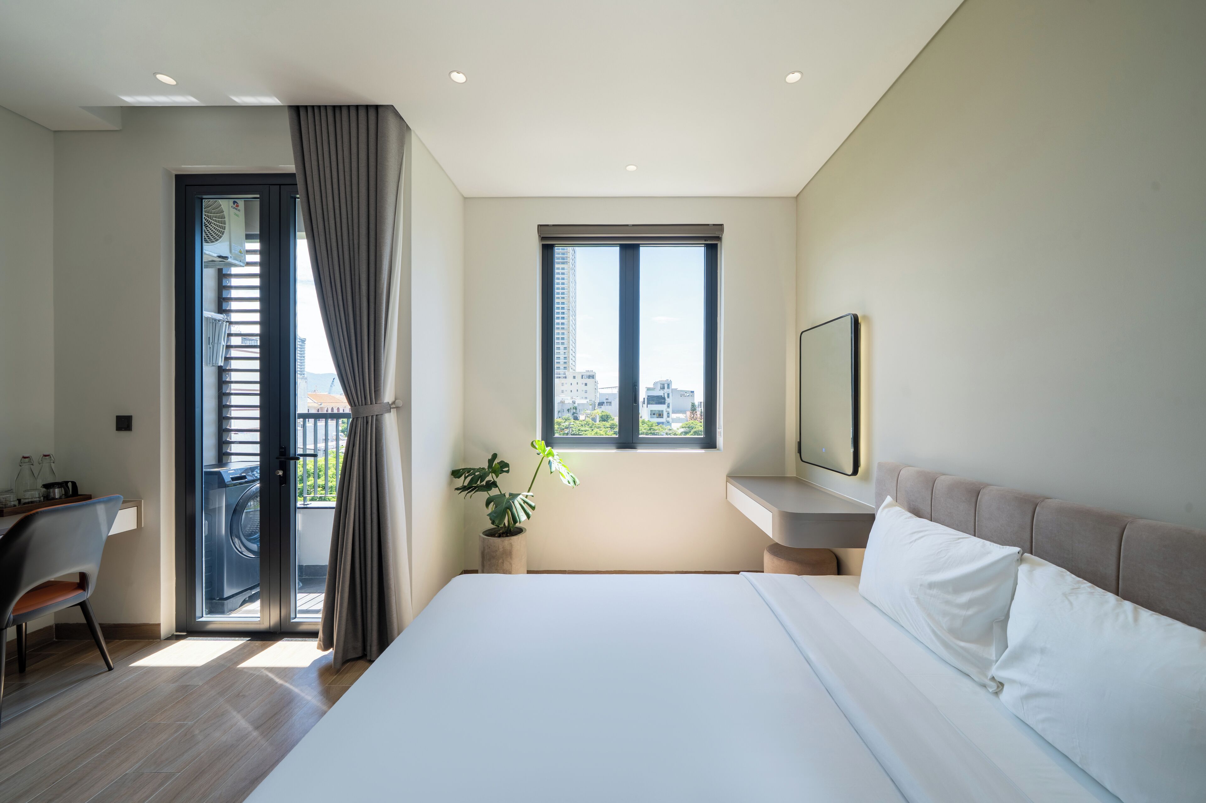 A modern hotel room with a bed, a desk, a chair, a potted plant, a window with a city view, and a balcony with a door.