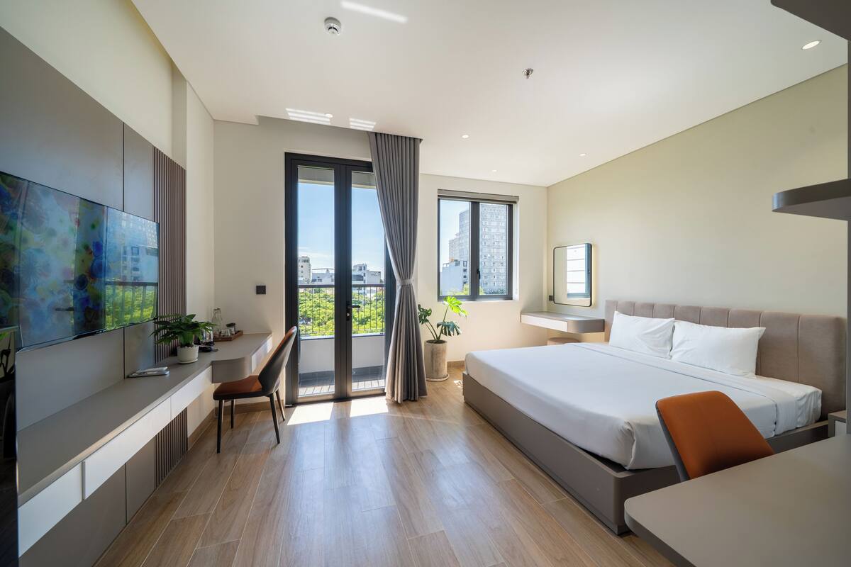 Habitación de hotel moderna con una cama grande, un escritorio, una silla, un televisor y un balcón con vistas a la ciudad.