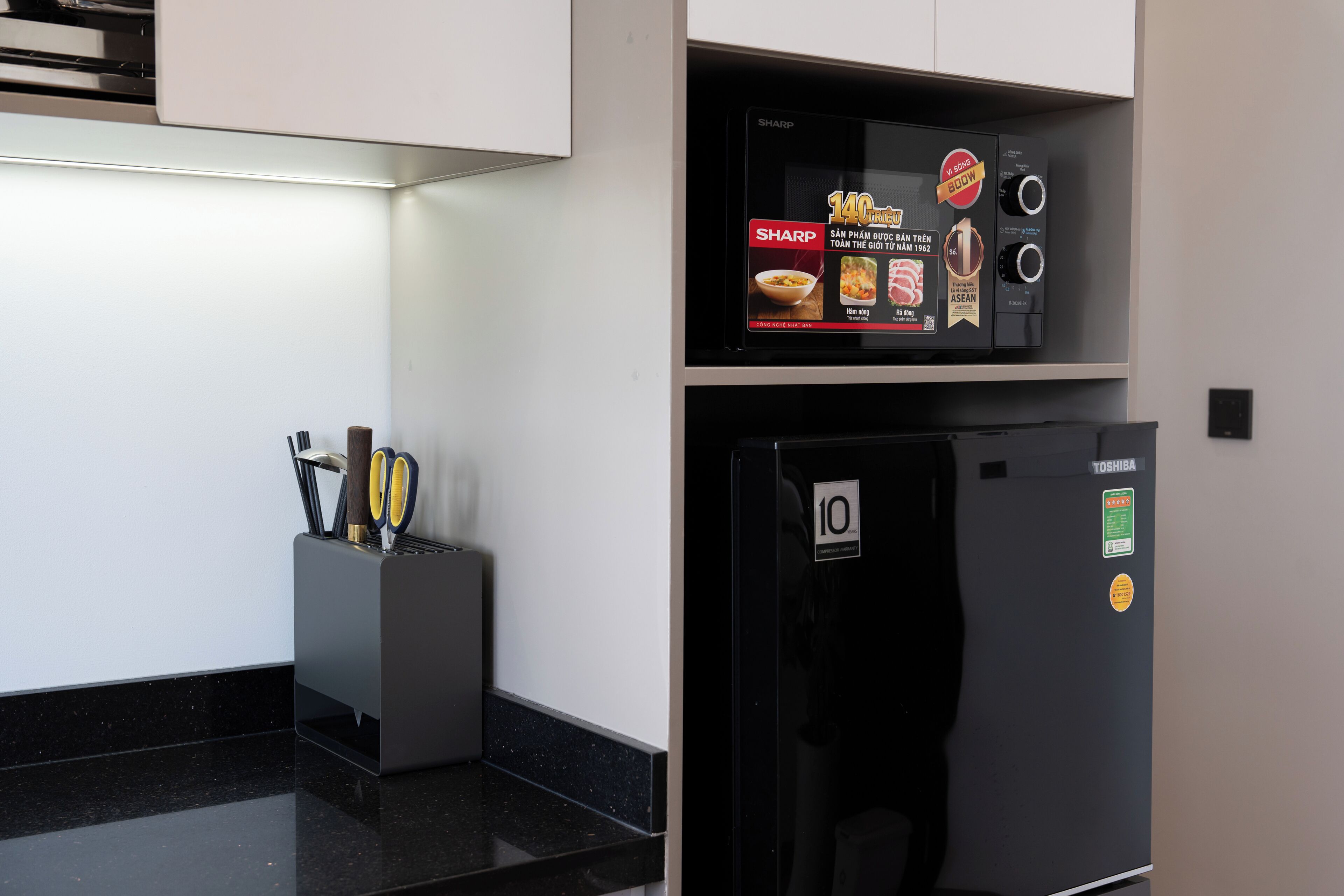 La cuisine est équipée d’un four à micro-ondes et d’un réfrigérateur, tous deux de couleur noire. On y trouve également un bloc à couteaux avec différents couteaux et une paire de ciseaux, posés sur le plan de travail.