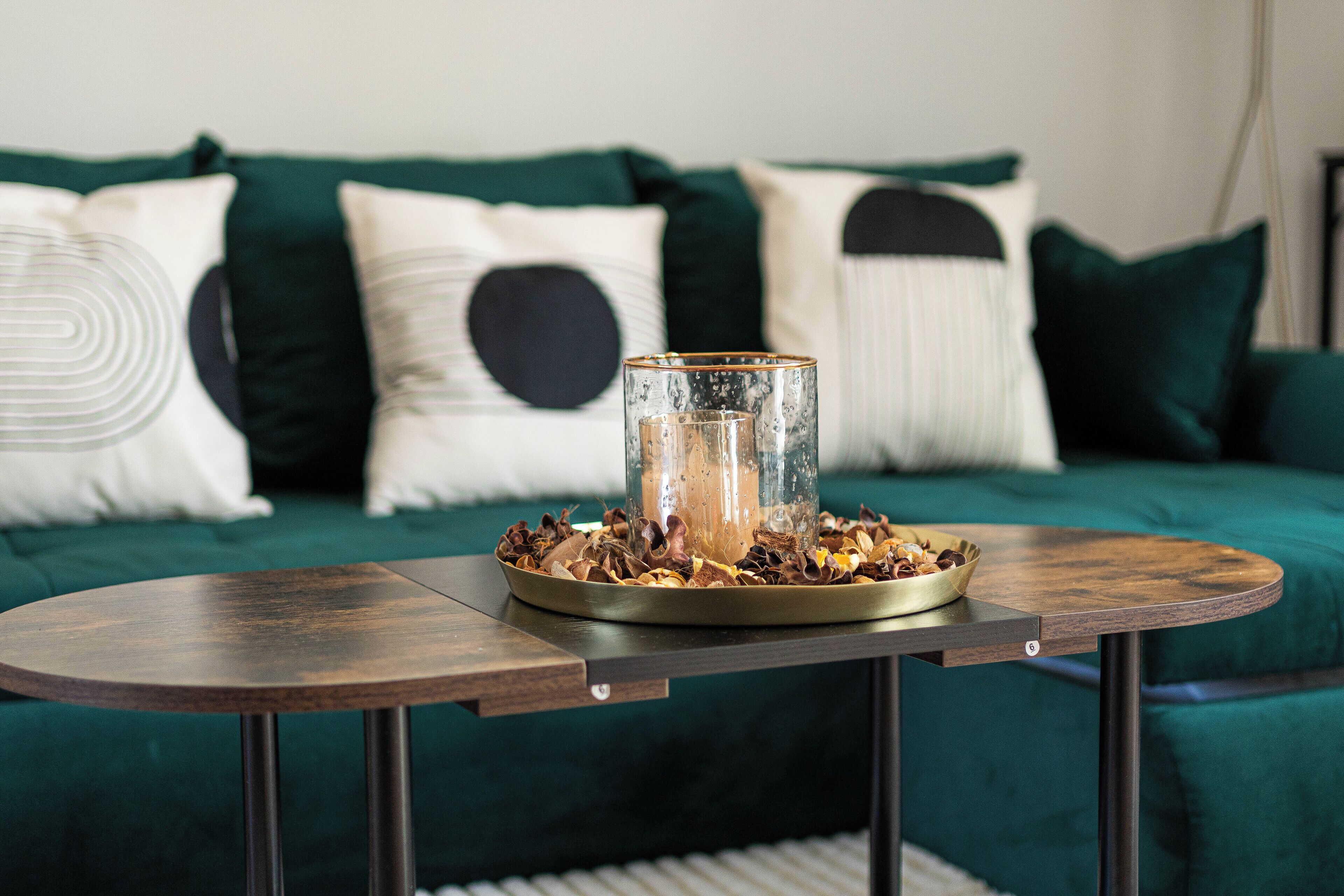 A round wooden table with a glass vase and dried flowers on a green sofa with patterned pillows.