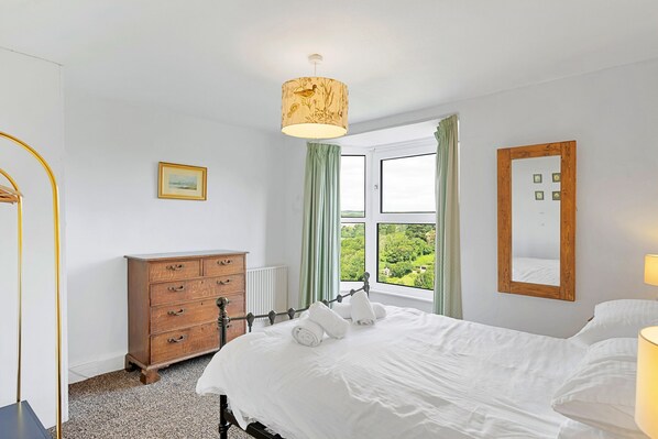 Quarto com cama, cómoda de madeira, radiador, janela com cortinas e candeeiro suspenso.