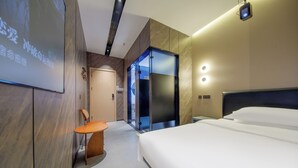 Une chambre d’hôtel moderne avec un grand lit, un bureau et une chaise. Une porte donne accès à une salle de bain et un ascenseur en verre est également présent.