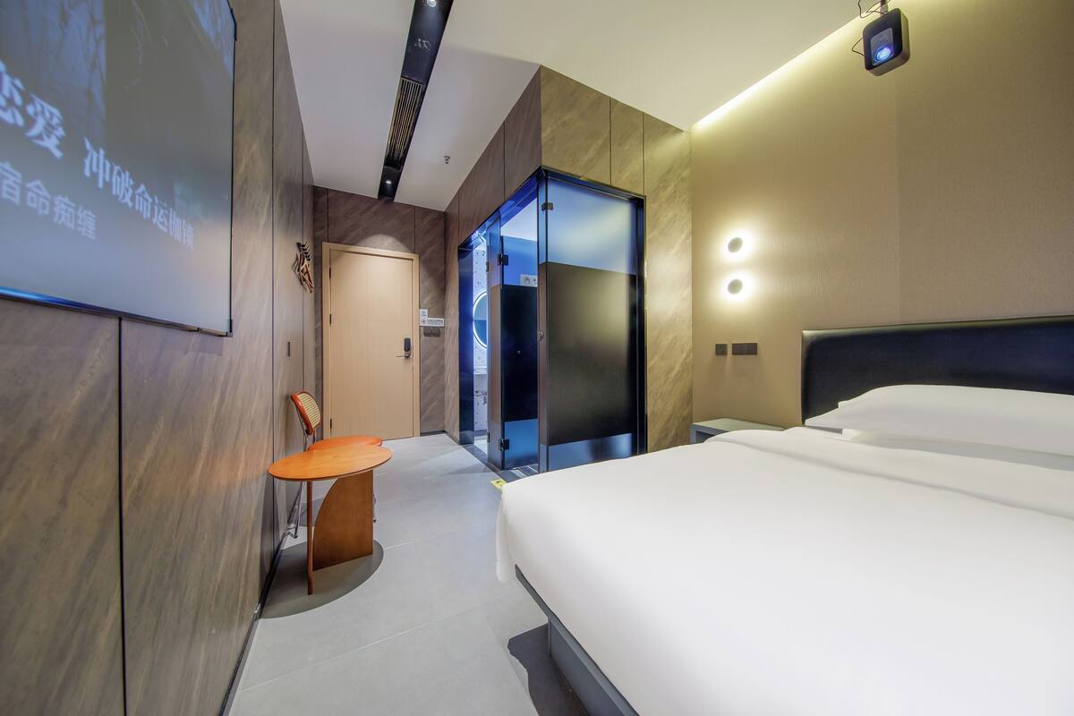 Une chambre d’hôtel moderne avec un grand lit, un bureau et une chaise. Une porte donne accès à une salle de bain et un ascenseur en verre est également présent.