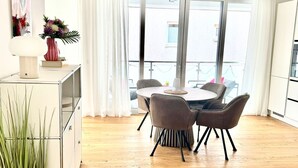 A modern living room with a dining area, wooden flooring, a white cabinet, a potted plant, and a large window with curtains. - "Discover Comfort and Relaxation at Strandsegler Heiligenhafen!" (Heiligenhafen)