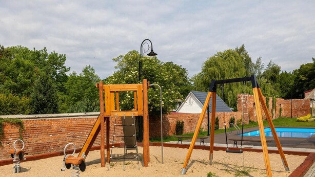 Un parque infantil con columpios y tobogán, rodeado por un muro de ladrillo y una piscina.