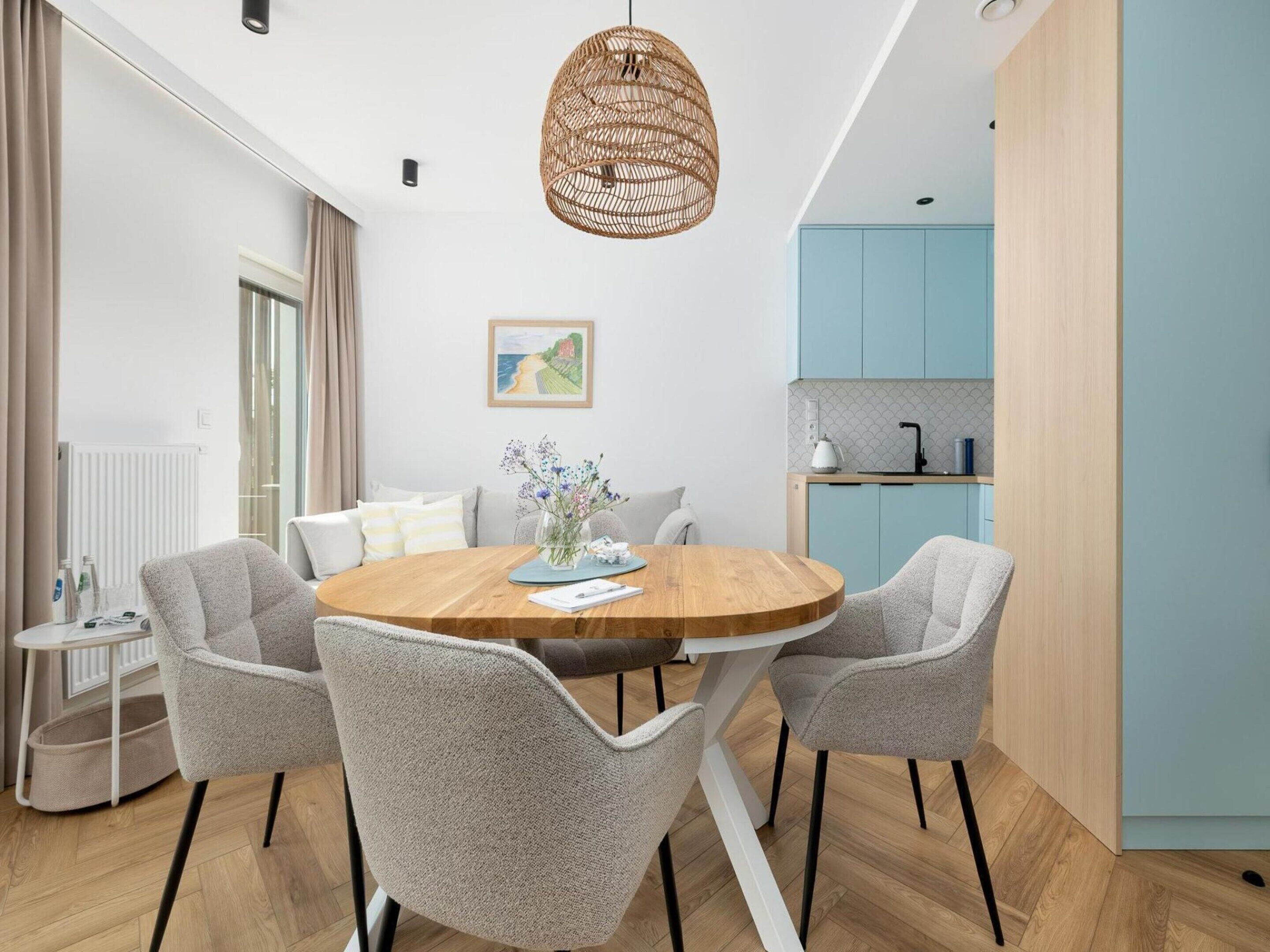 Un comedor moderno con una mesa redonda de madera, sillas grises tapizadas, una lámpara colgante de fibra natural y una cocina con armarios azul claro al fondo.