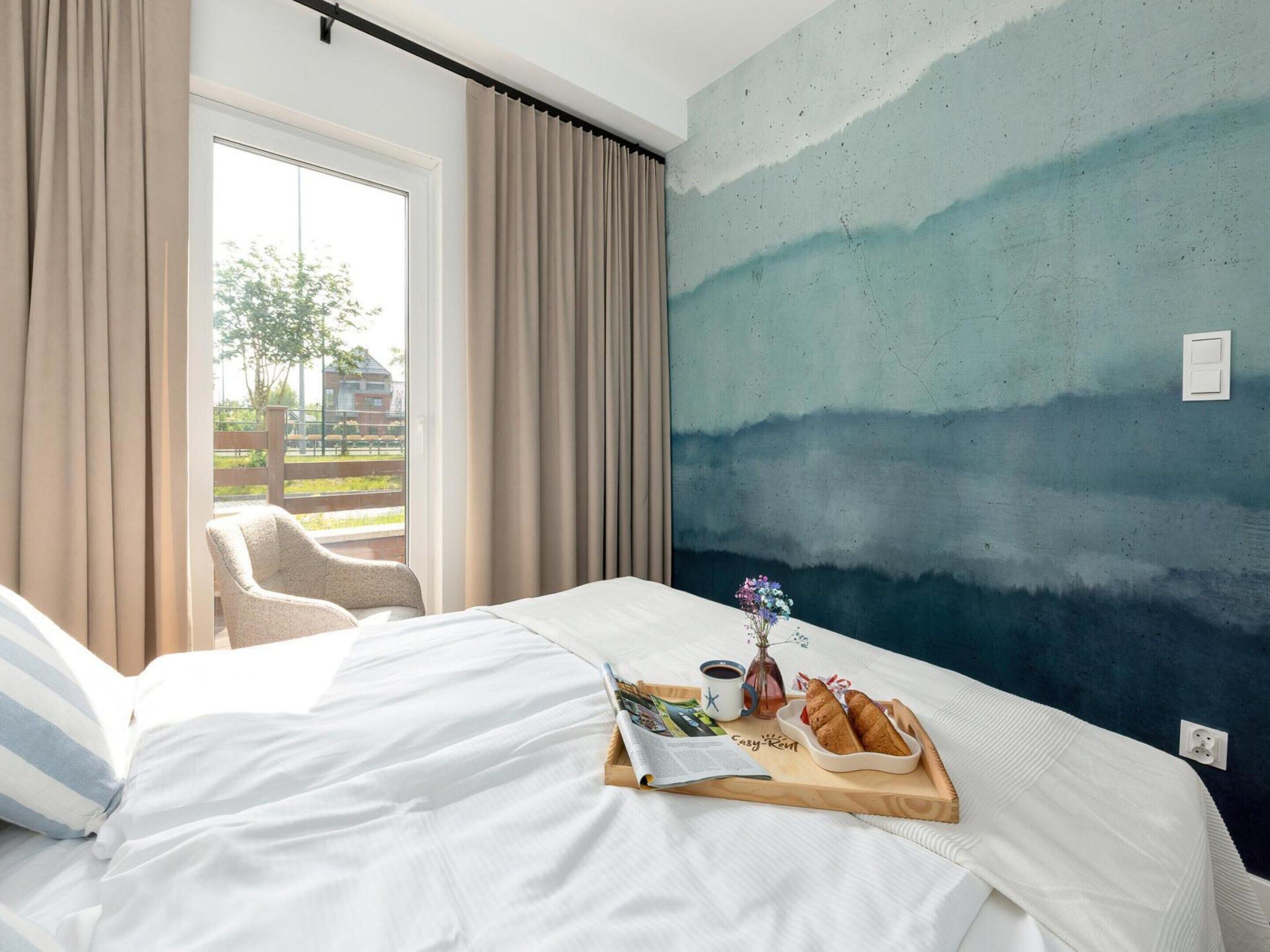 Une chambre d’hôtel avec un lit, une chaise, un plateau de petit-déjeuner avec des croissants, une tasse de café et un vase de fleurs.