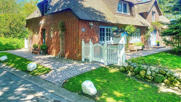 Ein traditionelles Haus mit Strohgedecktem Dach, einer Ziegel-Fassade, einem weißen Zaun und einem gepflegten Garten mit einer Steinmauer und großen Steinen.