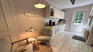 Eine Küche mit weißen Schränken, einer Kaffeemaschine und einer Mikrowelle. In der Ecke steht ein Stuhl und eine Lampe.