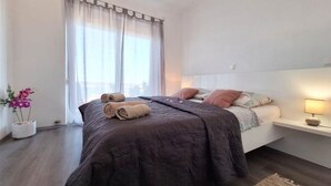Camera da letto con un letto, cuscini, un comodino, una lampada, una finestra con tende e una pianta in vaso.