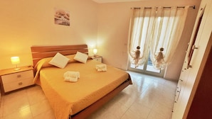 Une chambre d’hôtel comprenant un lit, deux tables de chevet, une commode, une fenêtre avec des rideaux légers et une porte.
