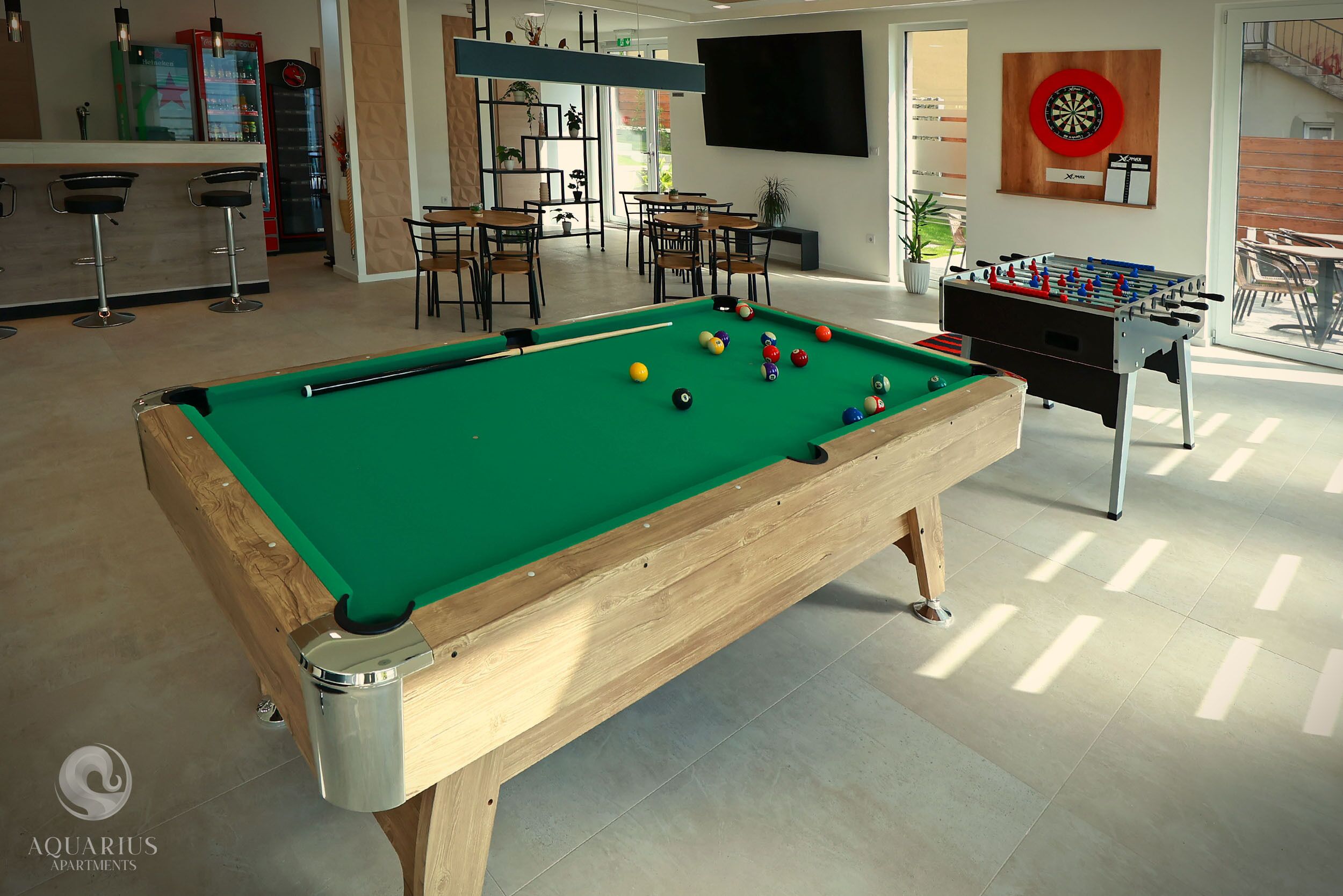 Ein Billardtisch mit Kugeln, ein Kicker und eine Dartscheibe in einem gut beleuchteten Innenraum mit Tageslicht.