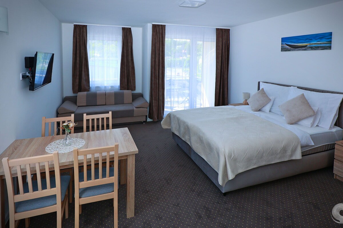 A hotel room with a bed, a sofa, a dining table with chairs, a TV, and a painting on the wall.