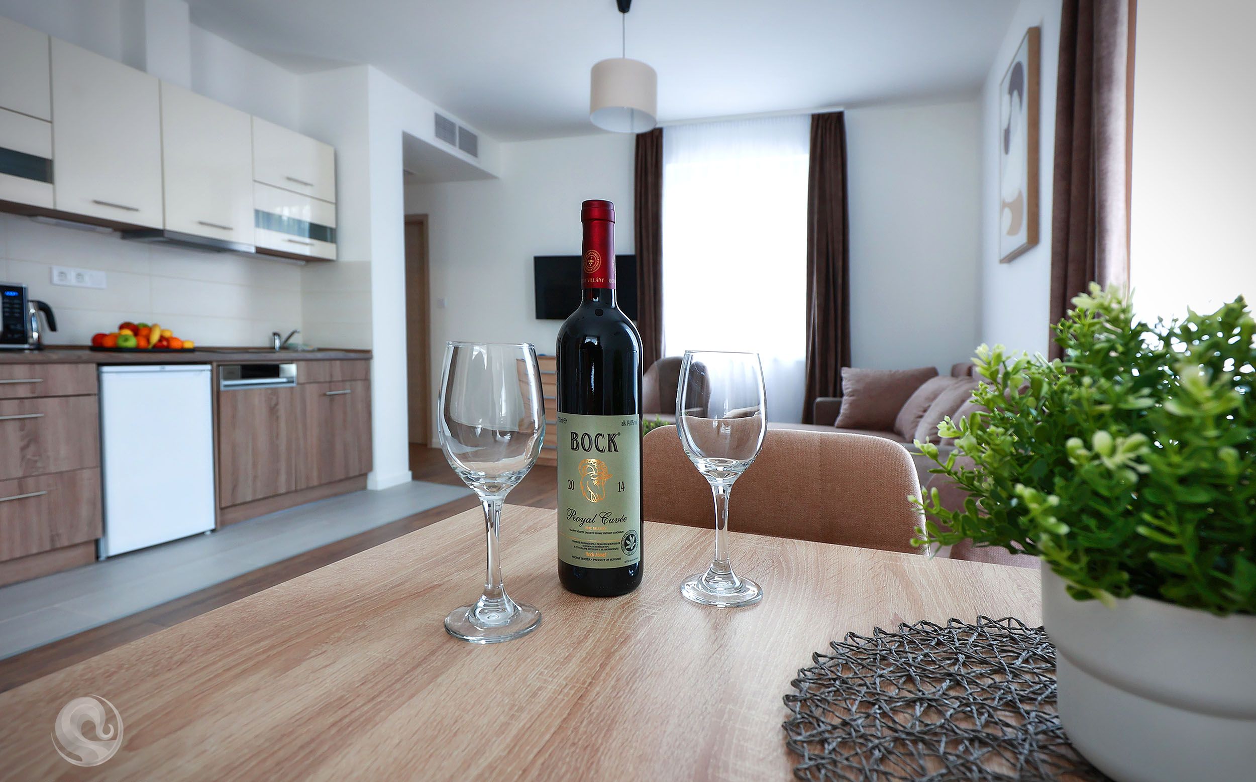 A modern kitchen with a wine bottle and two glasses on a wooden table, a potted plant, and a sofa in the background.