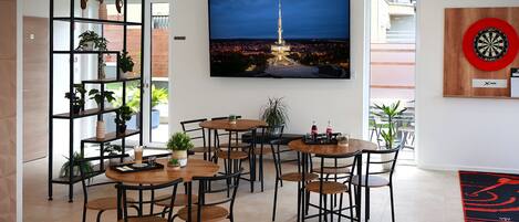 A modern dining area with multiple tables and chairs, a large TV screen displaying a cityscape, and a dartboard on the wall.