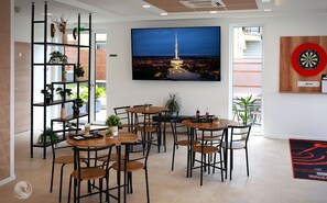 A modern dining area with multiple tables and chairs, a large TV screen displaying a cityscape, and a dartboard on the wall. - Aquarius Apartments (Miskolc)