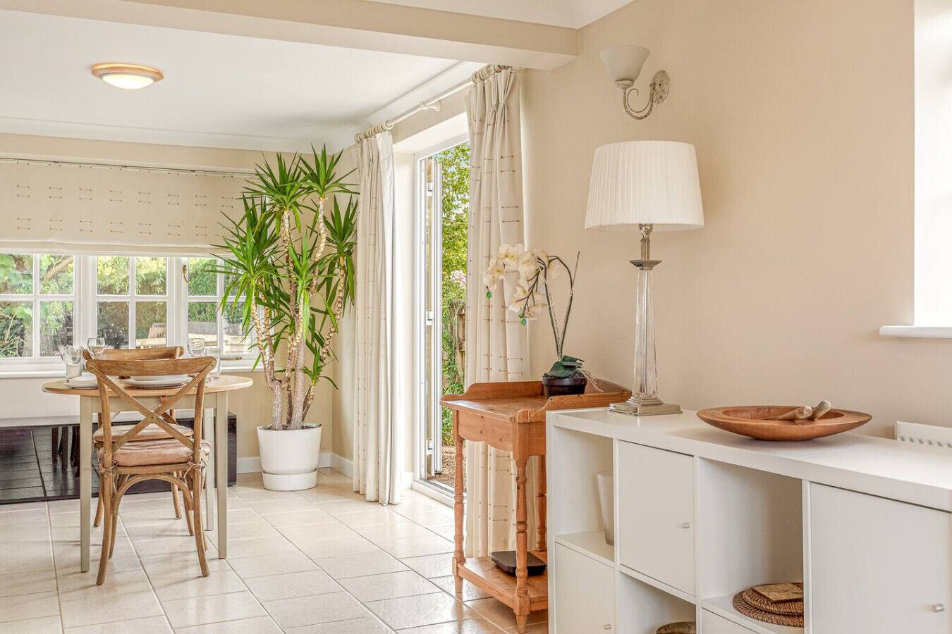 Un comedor luminoso con una mesa y sillas de madera, plantas en macetas y un armario blanco con una lámpara.