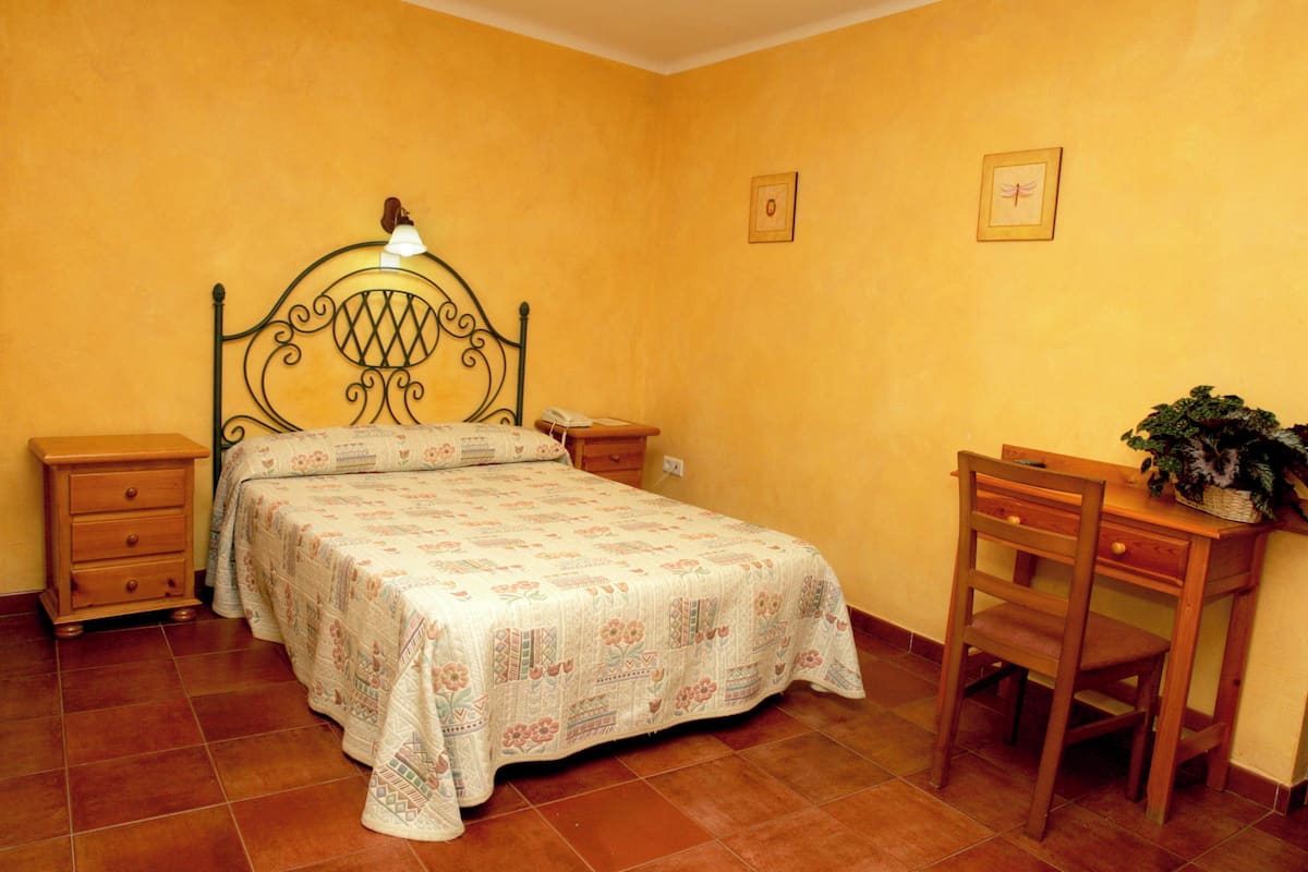 A bedroom with a bed, wooden nightstands, a desk with a chair, and a potted plant.