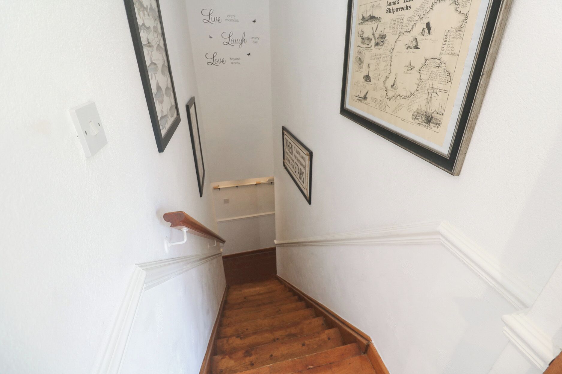 Un escalier avec une rampe en bois et un mur blanc orné de tableaux encadrés et d’une carte.