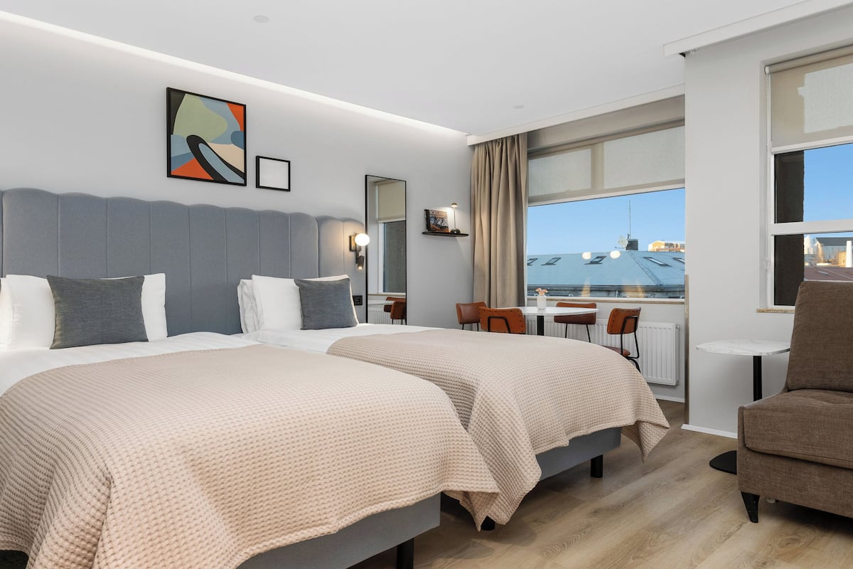 Habitación de hotel con dos camas, una zona de estar con una mesa y sillas, un ventanal con vistas a la ciudad y un cuadro en la pared.