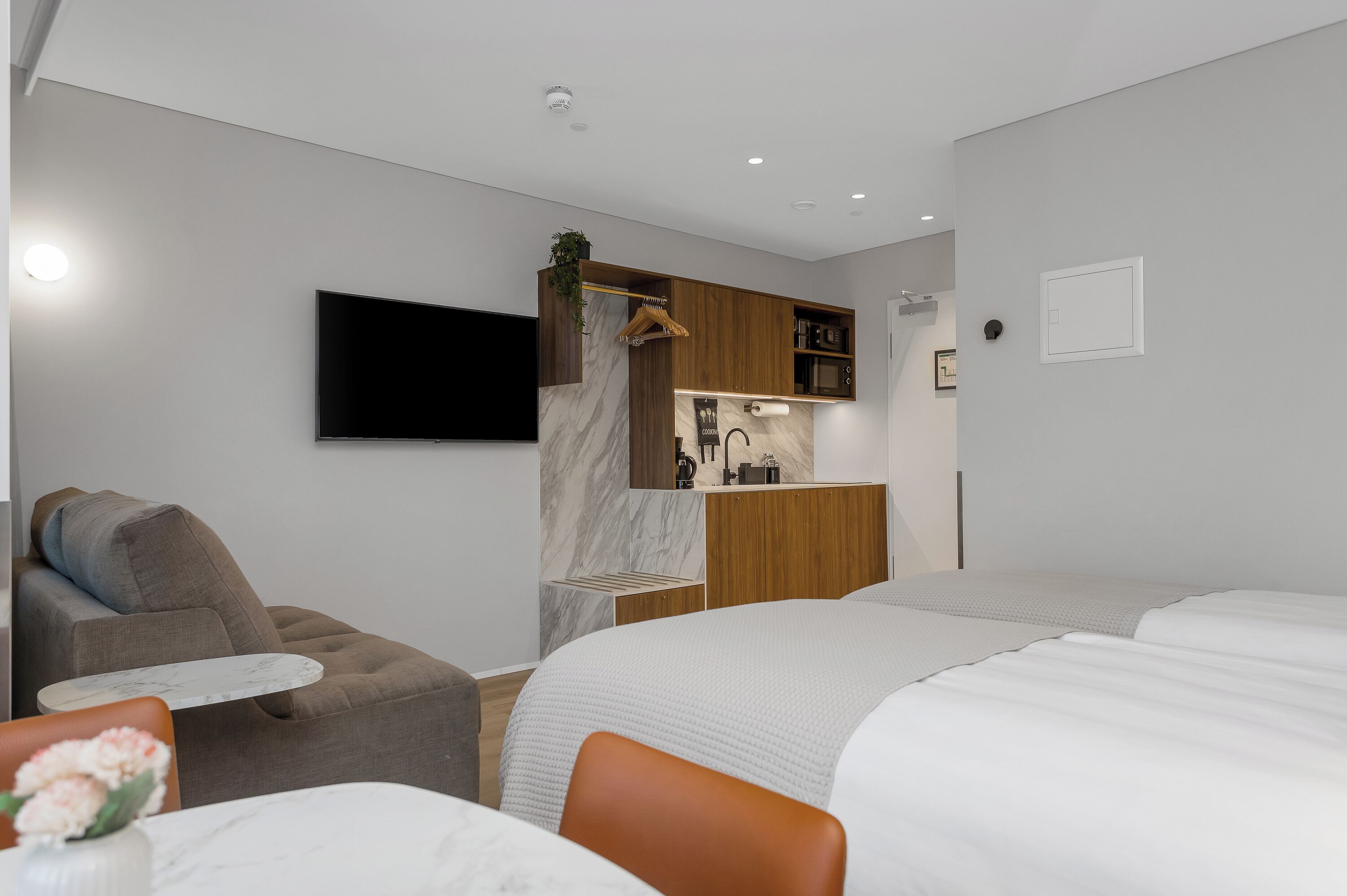 Moderni hotellihuone, jossa on sänky, sohva, pieni pöytä, televisio, keittiöalue ja amppelissa roikkuva kasvi.