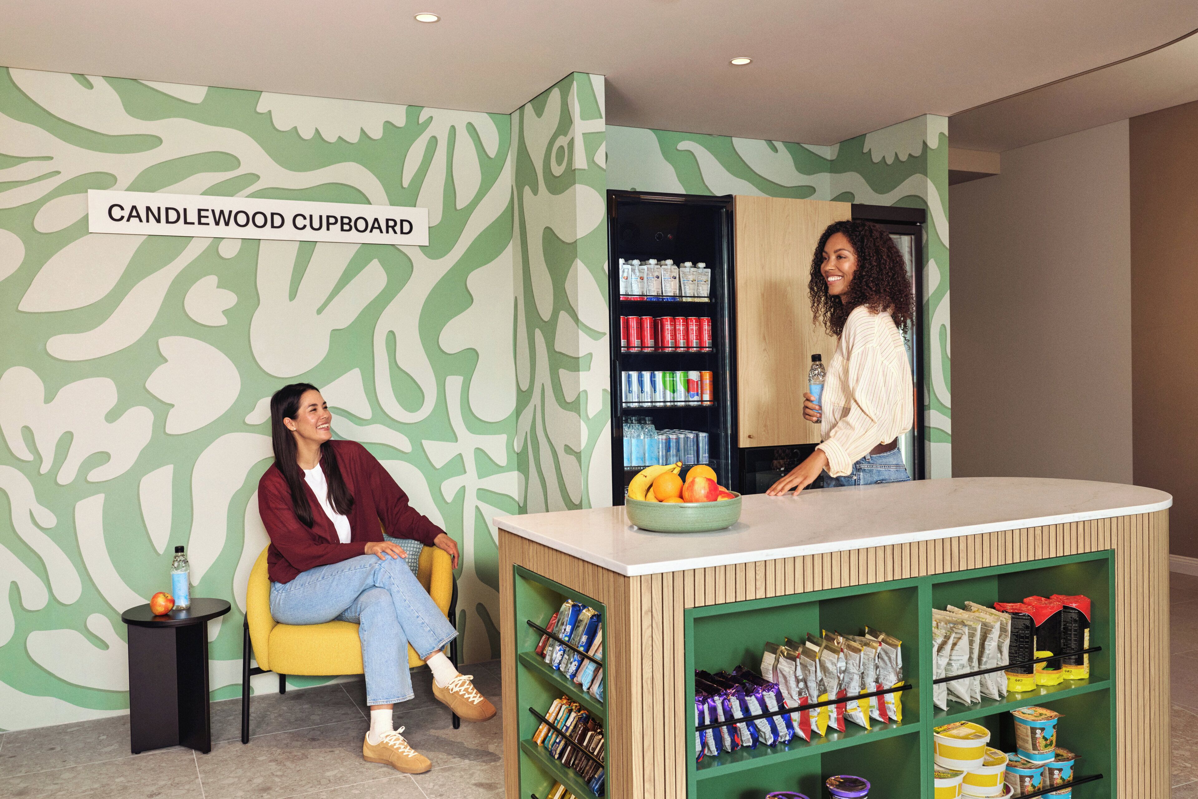 A room with a green patterned wall, a yellow chair, a white table, and a shelf with snacks and drinks.