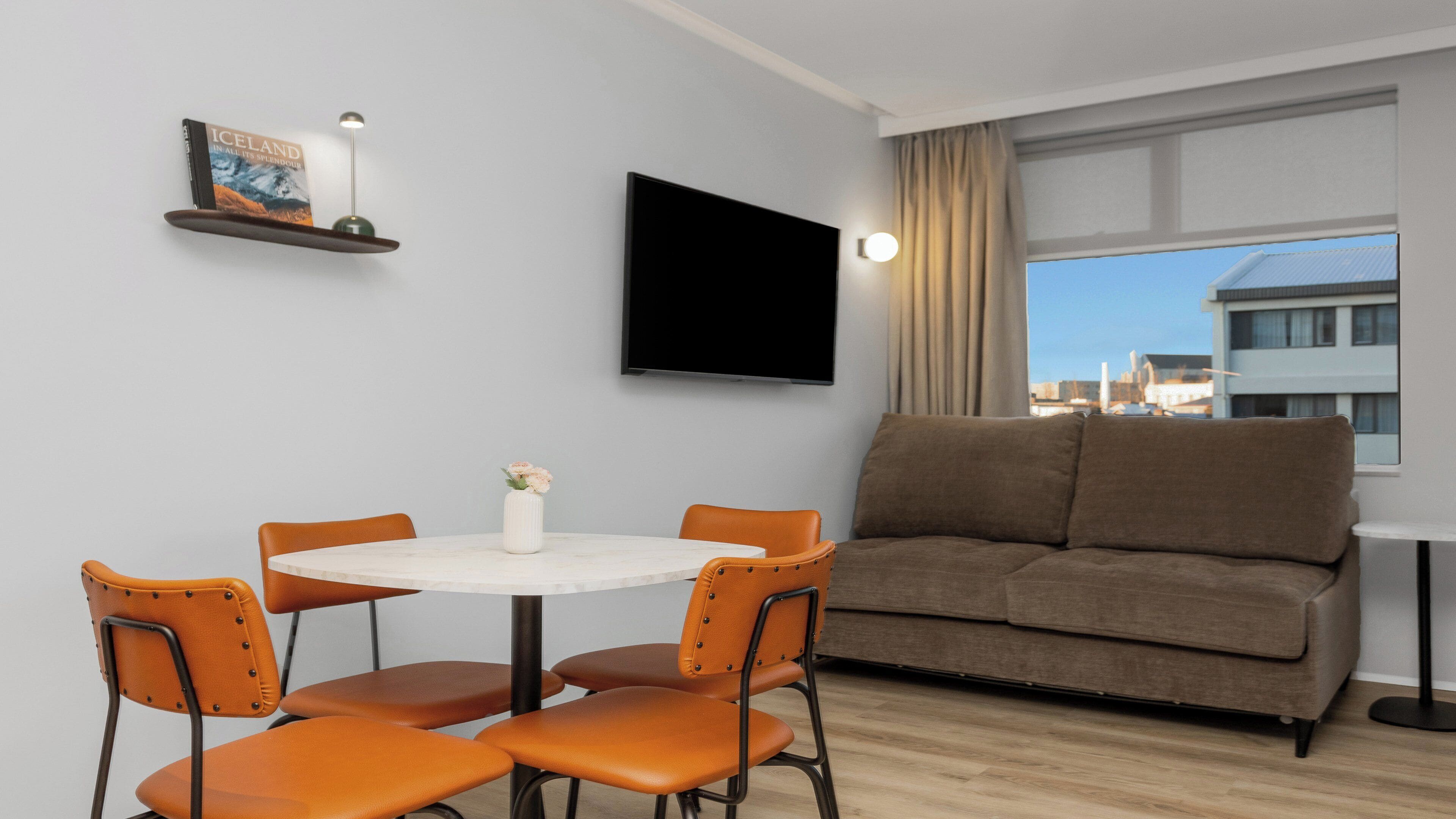 Una sala de estar moderna con un sofá, una mesa de comedor con sillas, un televisor de pantalla plana montado en la pared y una ventana con vista a la ciudad.