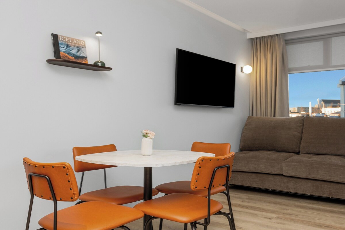 Una sala de estar moderna con un sofá, una mesa de comedor con sillas, un televisor de pantalla plana montado en la pared y una ventana con vista a la ciudad.