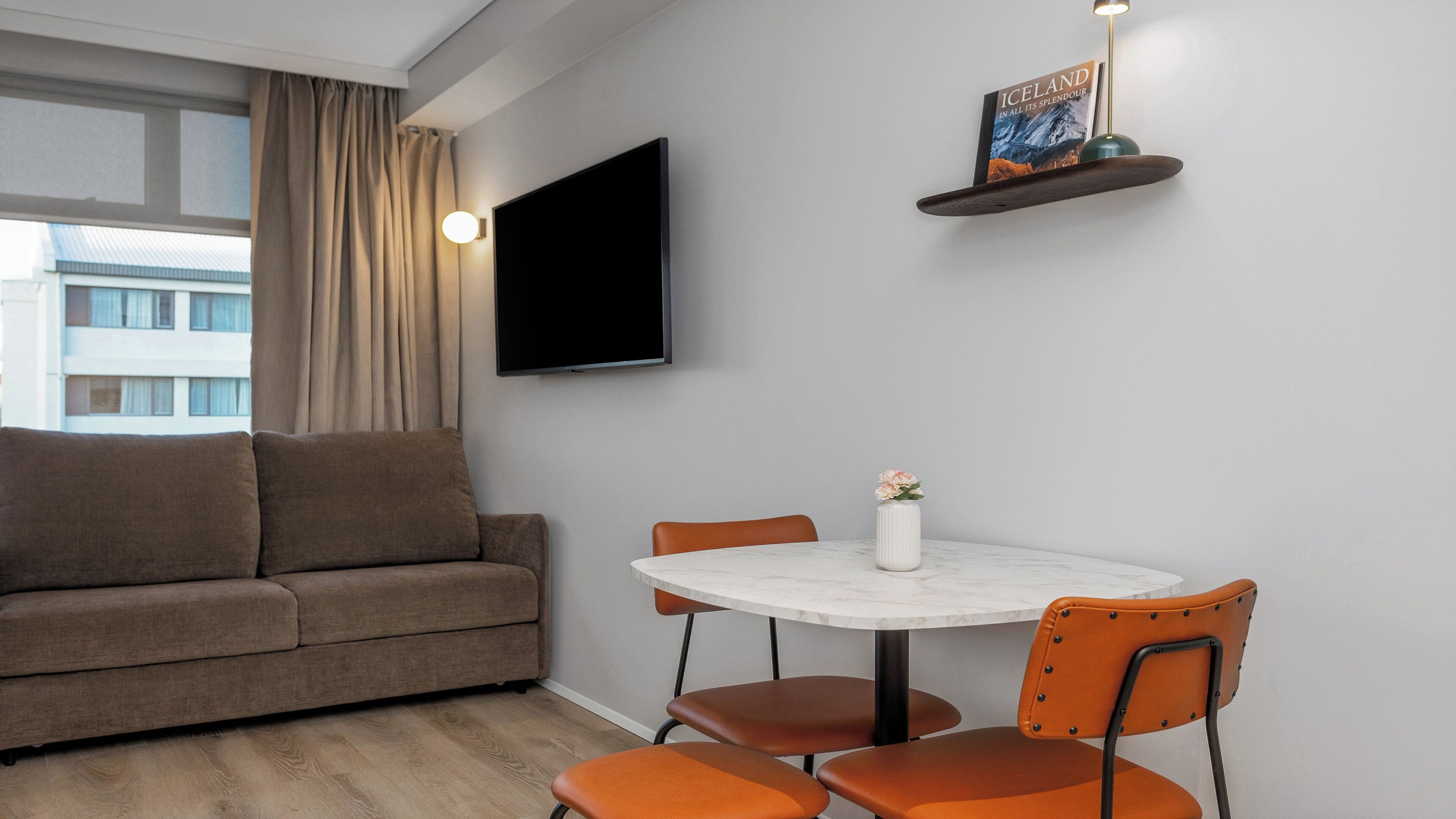 Un salon moderne avec un canapé, une table ronde et deux chaises. Un téléviseur à écran plat est fixé au mur, et un livre intitulé « ISLANDE » est exposé sur une étagère.