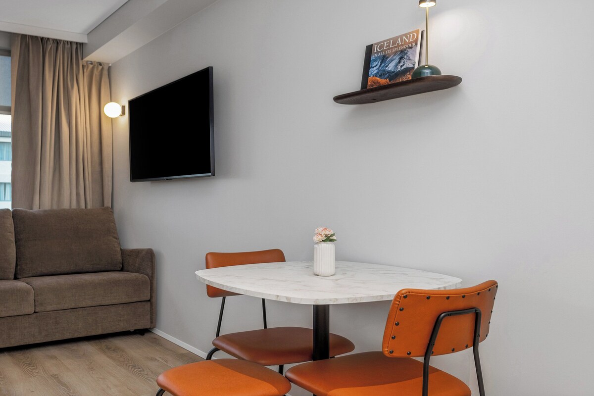 Una sala moderna con un sofá, una mesa redonda y dos sillas. Un televisor de pantalla plana está montado en la pared, y un libro titulado 