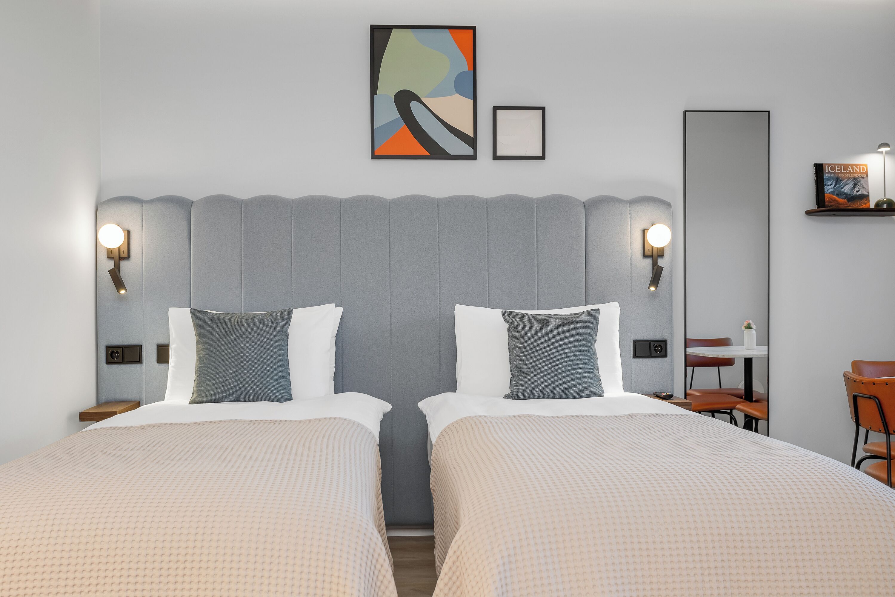 A hotel room with two beds, a desk, a chair, and a bookshelf.