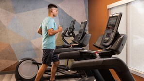 Una persona usando una cinta de correr en un gimnasio con un diseño geométrico en la pared y una ventana con persianas.
