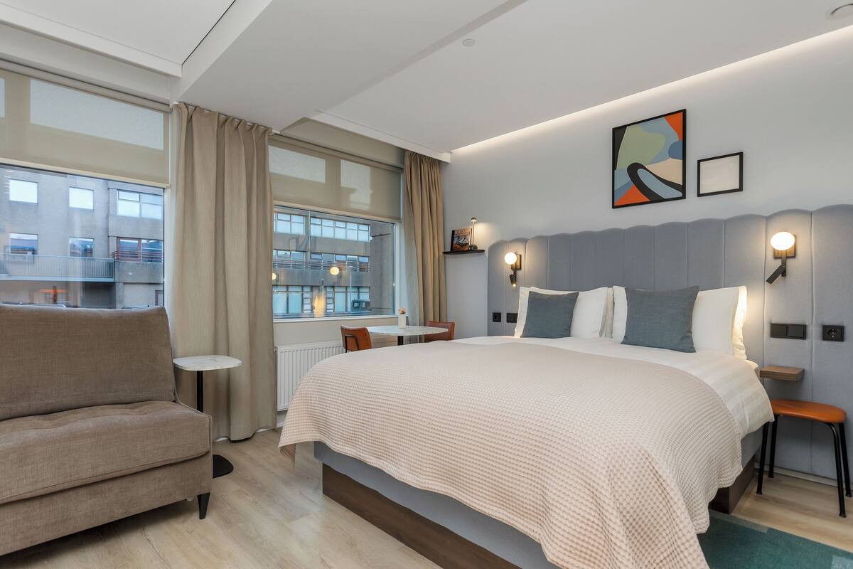 Un hotel moderno con una cama grande, un sof á, una mesita y vistas a la ciudad a través de amplias ventanas.