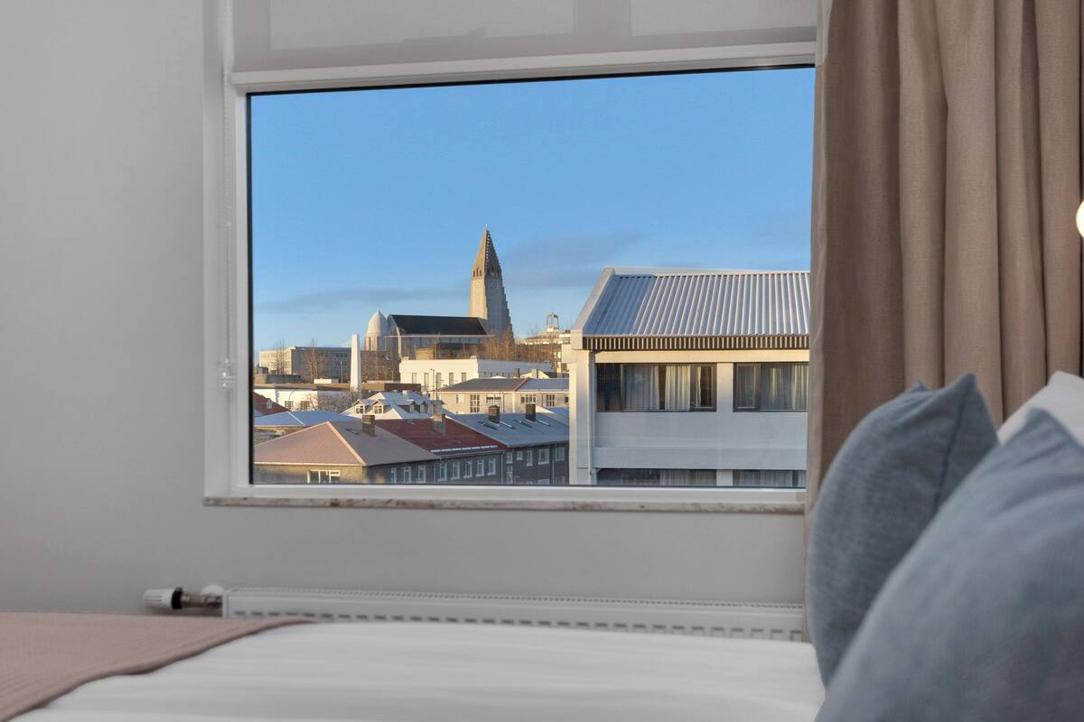 Habitación de hotel con cama, ventana y vista a un paisaje urbano que incluye una iglesia y edificios.