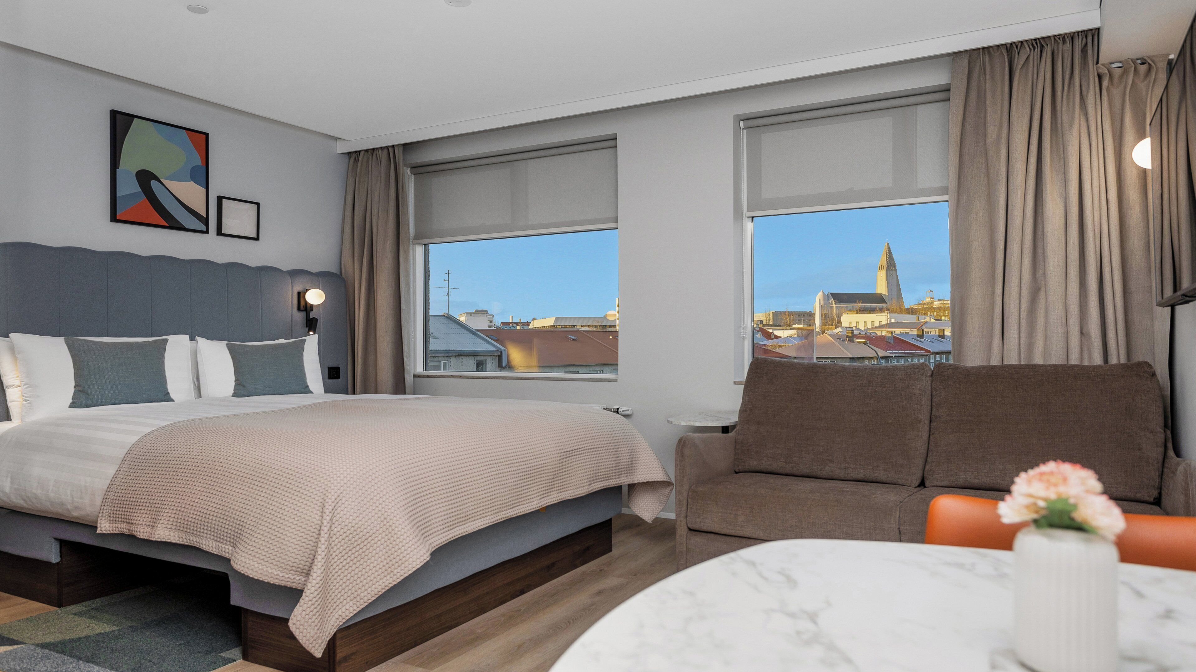 A modern hotel room with a large bed, a sofa, a dining table, and a view of a cityscape.