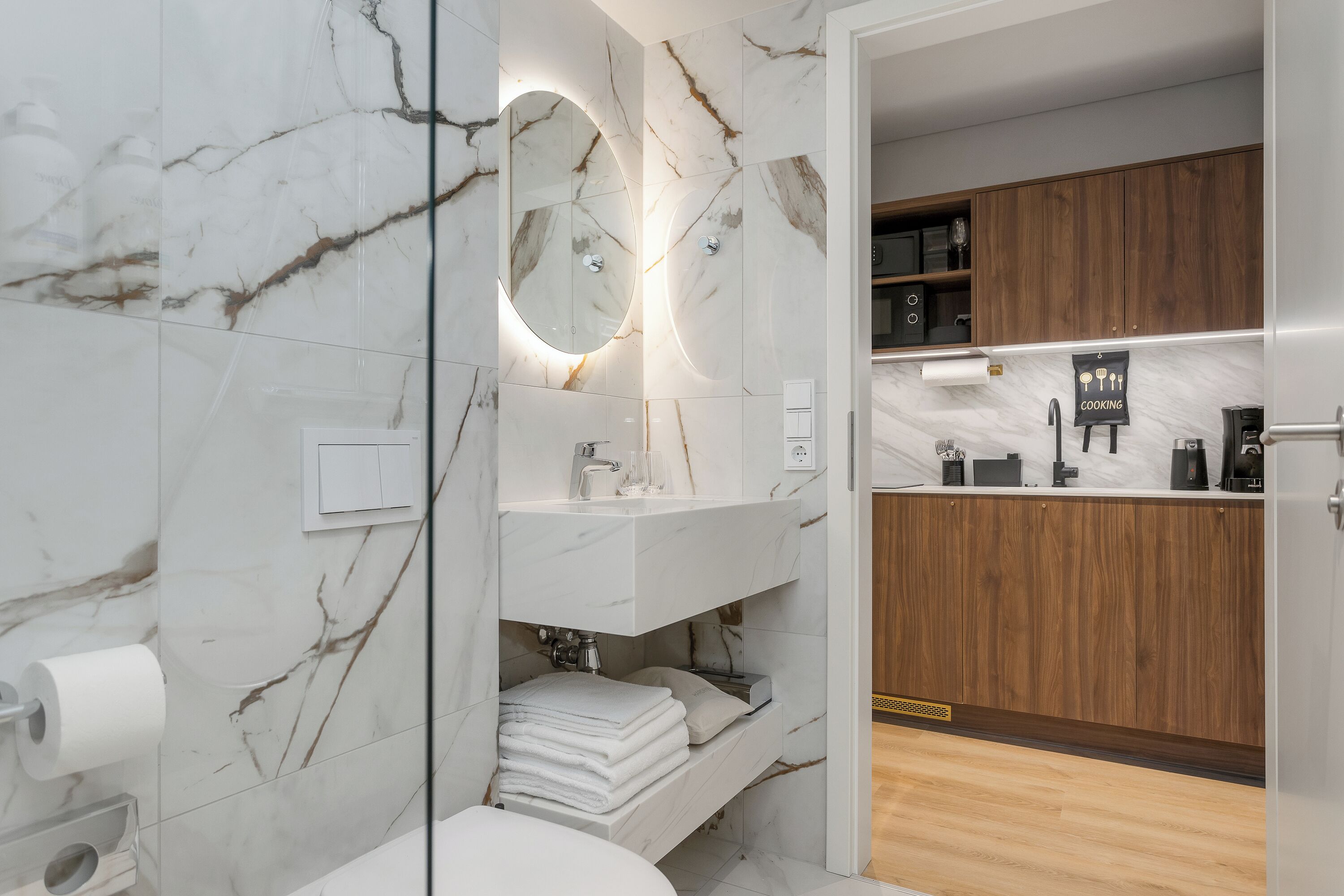 Ein modernes Badezimmer mit Marmorwänden, einem Waschtisch mit Spiegel und einem Holzkorpus in der angrenzenden Küche.