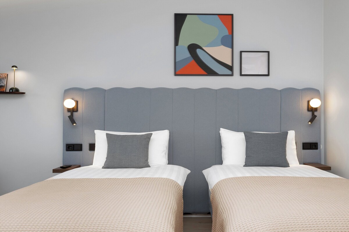 Habitación doble con una cama y cabecera gris, dos mesitas de noche y una lámpara fijada a la pared.