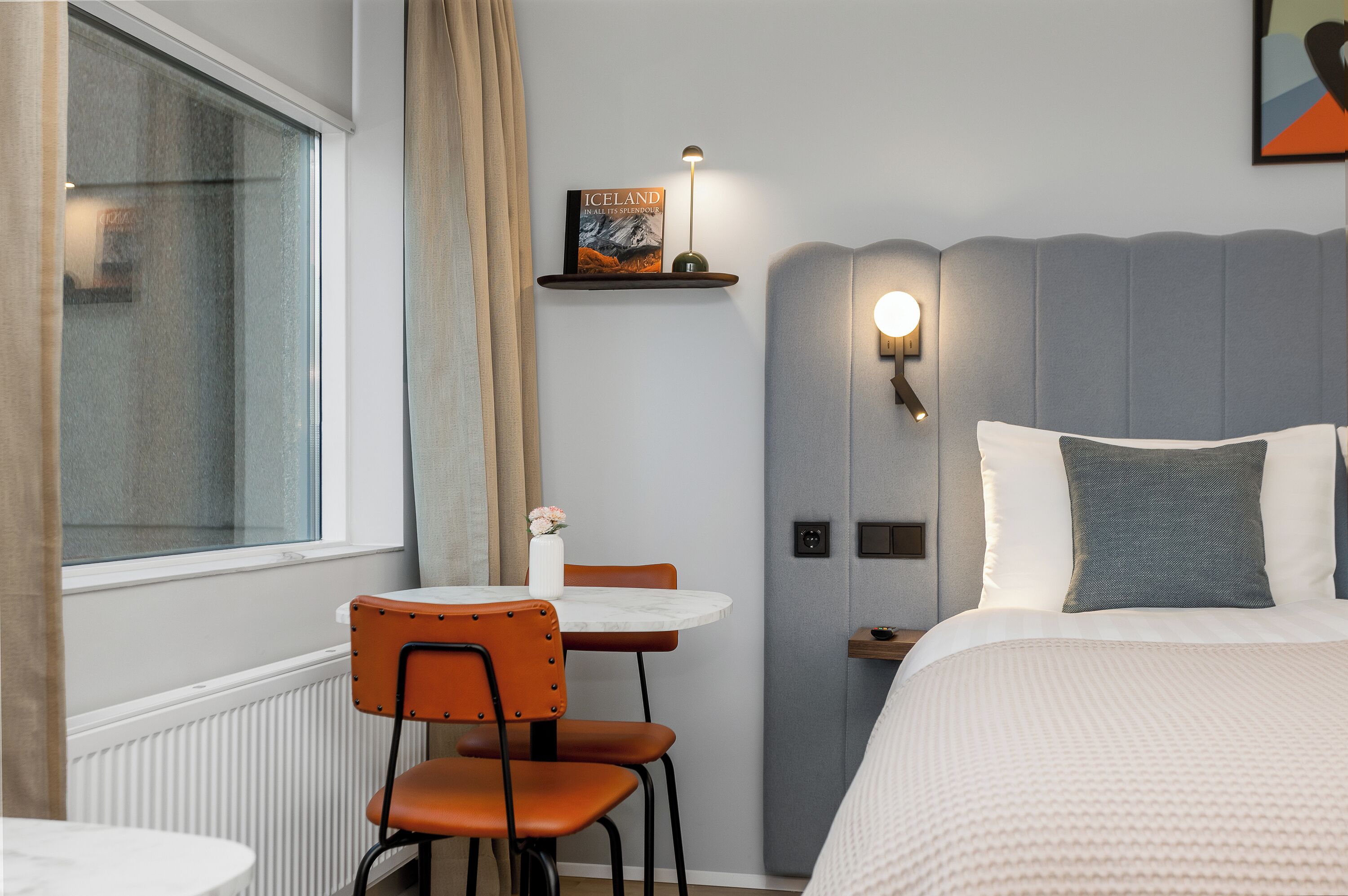 A hotel room with a bed, a bedside table, a chair, a small table with a vase, a book on a shelf, and a window with curtains.