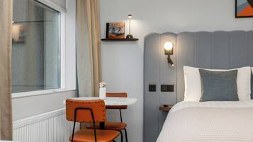 A hotel room with a bed, a bedside table, a chair, a small table with a vase, a book on a shelf, and a window with curtains.